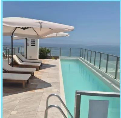 a swimming pool with an umbrella and chairs and the ocean at Departamento frente al mar costa verde in Lima